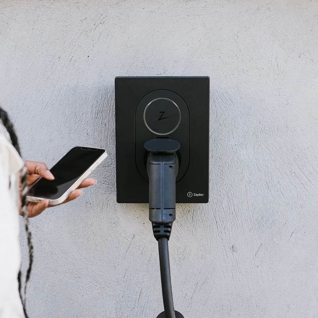 Zaptec Go -latausasema asennettuna harmaaseen betoniseinään. Nainen seisoo latausaseman vieressä ja käyttää Zaptec Portal -sovellusta latausasemaa ohjaten