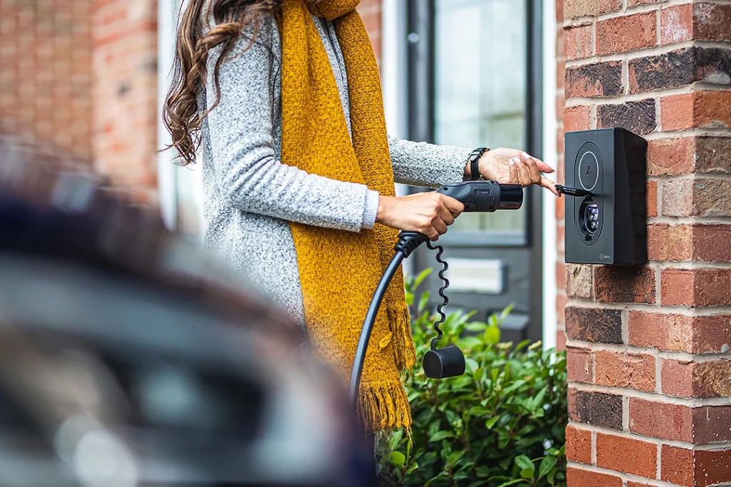 Nainen asettamassa Zaptec-latausasemaan sähköauton latauskaapelia. Latausasema on asennettuna ulkona punaiseen tiiliseinään.